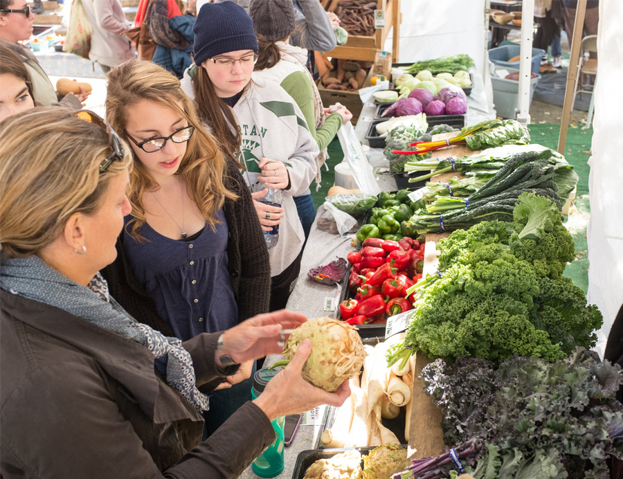 A crowd of people candidly stand in front of a table full of fresh vegetables for sale at the Burlington Farmers Market.