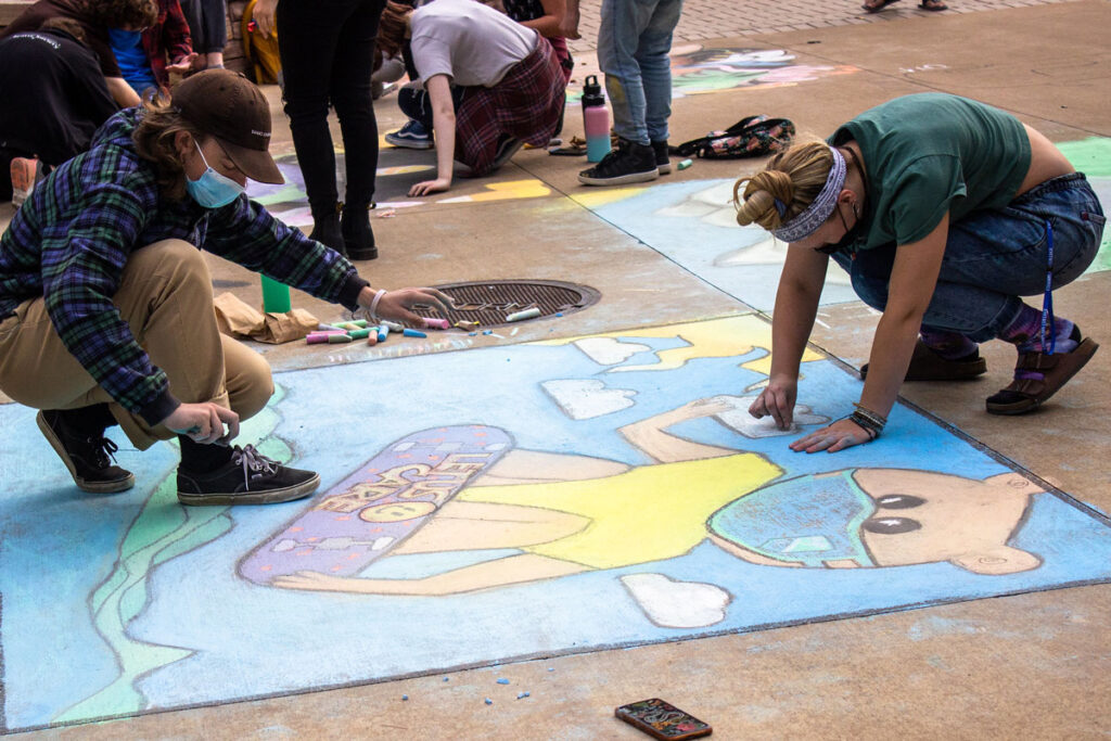 Two students draw Chauncey on a skateboard with sidewalk chalk