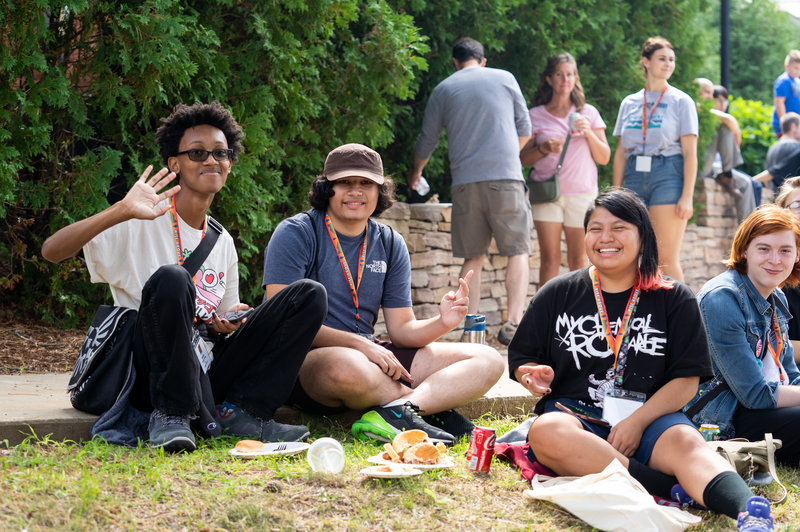 A group of four students eat lunch on the green and smile at the camera.