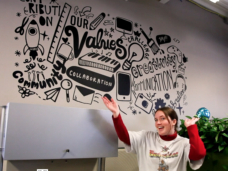 A student holds her hands up, pointing at a wall of artwork she created that displays a variety of words including "our values," "collaboration," "creativity," and more.