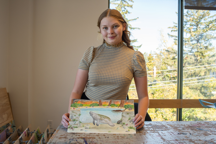 A student holds a piece of colorful artwork on a table.