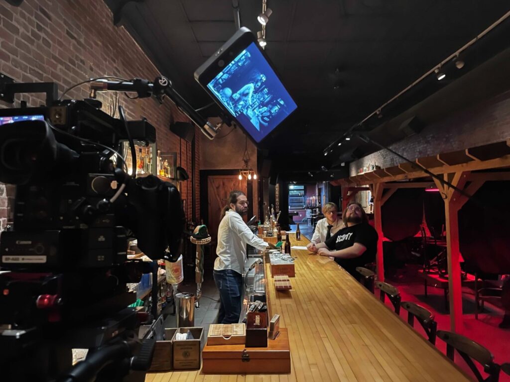 Three actors surrounding a bar; two patrons, one bartender. A camera is pointed at them.