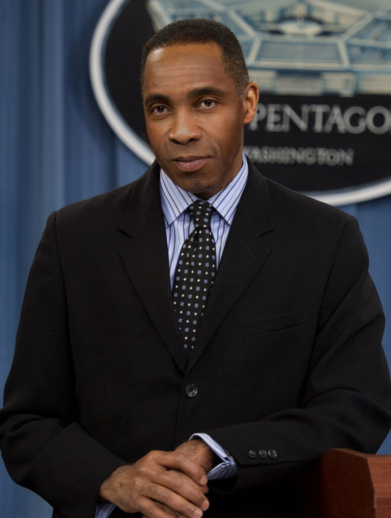 JJ Green wears a black suit and square checkered tie, standing in front of a Pentagon sign