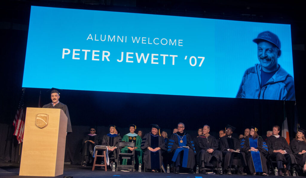 Peter Jewett stands at a wooden podium on stage in front of 25 people seated on stage, with a large blue screen above reading "alumni welcome, Peter Jewett '07"