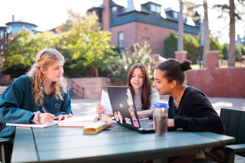 Three Champlain students sit around a table in the courtyard, looking at a sheet of paper.
