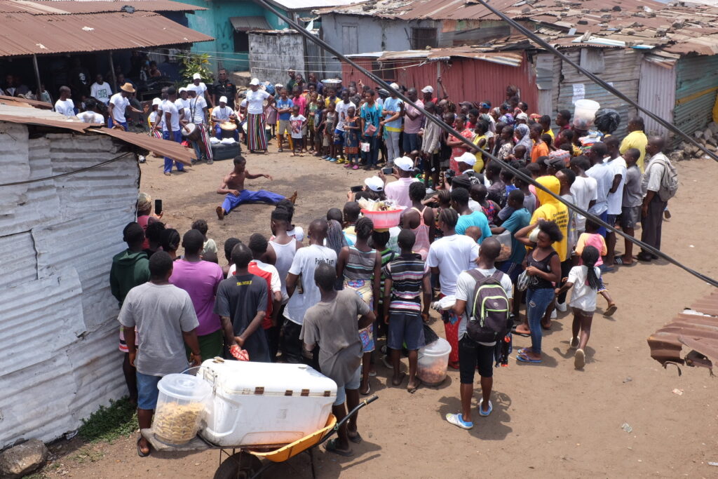 A significant crowd gathers around a theatrical performer on the streets in Liberia.